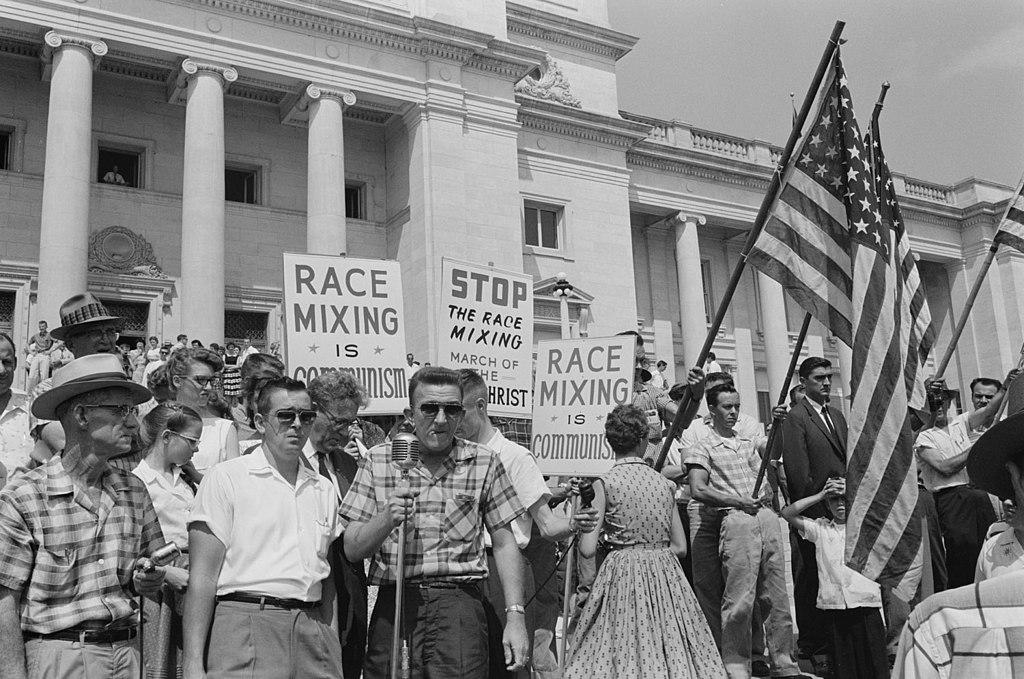Photo of anti-integration protestors holding signs reading "race mixing is communist" and "Stop the race mixing, march of the antichrist."